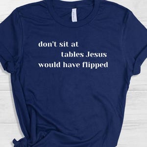 May include: Navy blue t-shirt with the white text "don't sit at tables Jesus would have flipped". The shirt is made of a soft material and has short sleeves. The shirt is a casual wear item.