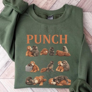 May include: Green sweatshirt with the word "PUNCH" in orange block letters. The sweatshirt features a repeating pattern of cartoon monkeys in various poses. The monkeys are in shades of brown and gray.
