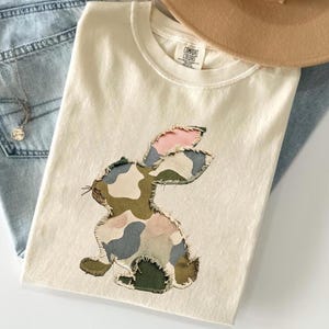 May include: Cream-colored t-shirt featuring a camouflage bunny applique with pink and green accents. The shirt is folded, with a pair of blue jeans and a tan hat in the background. A silver necklace with a pendant is also visible.