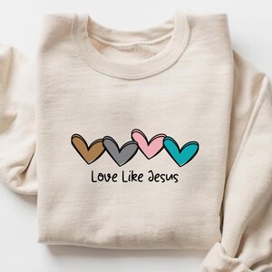 May include: A cream-colored sweatshirt featuring a row of four outlined hearts in brown, gray, pink, and teal. The phrase "Love Like Jesus" is printed in black below the hearts. The sweatshirt has a crew neck and long sleeves.