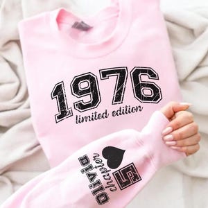 May include: A light pink sweatshirt with the year "1976" printed in black, along with "limited edition". The sleeve displays "Olivia Chapter 5" with a heart symbol. The sweatshirt is folded on a light-colored fabric.