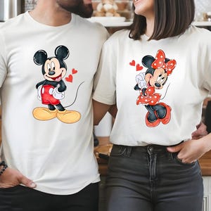 May include: Off-white t-shirts featuring Mickey and Minnie Mouse. Mickey is in red shorts and yellow shoes, with red hearts. Minnie is in a red polka-dot dress and bow, with red hearts. The shirts are a matching couple's set.