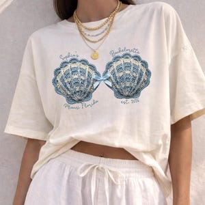 May include: Cream-colored t-shirt featuring a seashell design in blue and white crochet, with a blue bow. Text includes "Sophia's Bachelorette" and "Miami Florida est. 2026". The shirt is part of a bridal party theme.