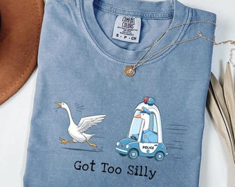 May include: A blue Comfort Colors t-shirt with a graphic of a goose being chased by a police car driven by a goose. The text "Got Too Silly" is printed below the graphic. A gold necklace is draped across the shirt.