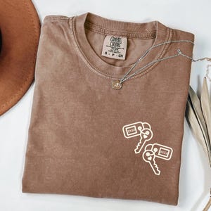 May include: A brown Comfort Colors t-shirt with a crew neck and short sleeves. The shirt features a white graphic of two keys with the numbers "1410" and "1221". A silver necklace with a small pendant is draped across the shirt.