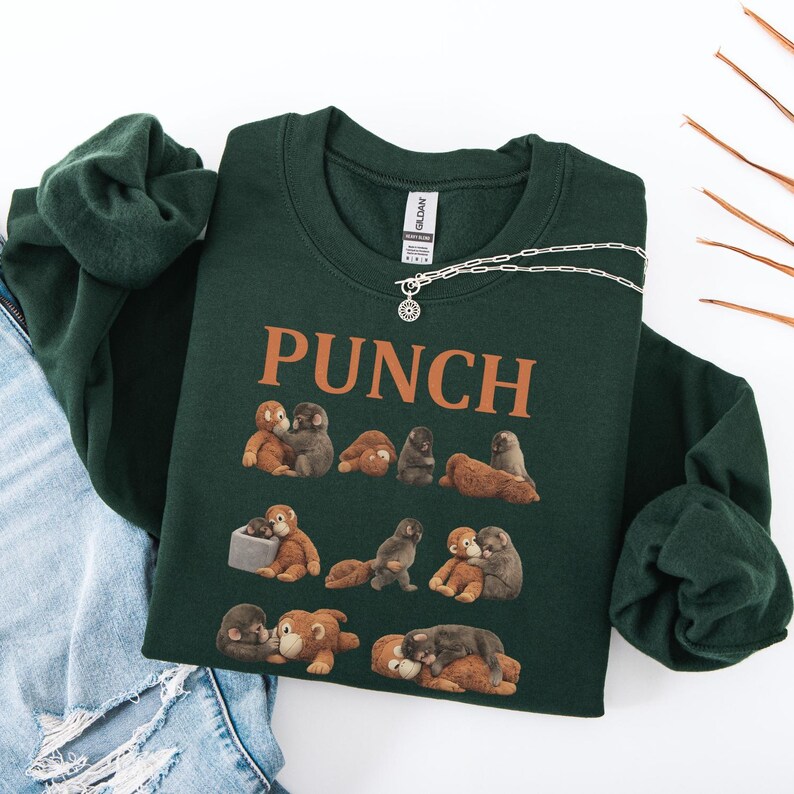 May include: A dark green sweatshirt with the word "PUNCH" in orange block letters. The sweatshirt features a pattern of small, cartoon-like monkeys in various poses. A silver necklace with a small pendant is draped on the sweatshirt.