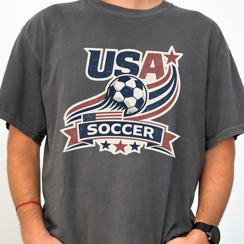 Pu&ograve; includere: Una t-shirt grigio antracite a maniche corte con una stampa grafica recante il testo USA SOCCER. Il design include un pallone da calcio, linee curve rosse e blu, una piccola bandiera americana e tre stelle nei colori rosso e blu.