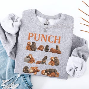 May include: A heather gray sweatshirt with the word "PUNCH" in orange block letters. The sweatshirt features a repeating pattern of cartoon monkeys in various poses. The sleeves are cuffed, and a silver necklace with a sunburst pendant is visible.