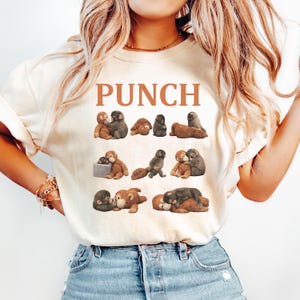 May include: Cream-colored t-shirt with the word "PUNCH" in brown block letters. The shirt features a repeating pattern of cartoon monkeys in various poses, in shades of brown and gray.
