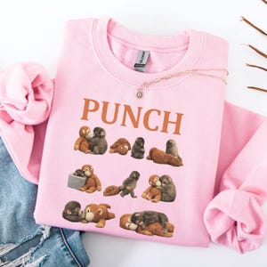 May include: A light pink sweatshirt with the word "PUNCH" in brown block letters. The sweatshirt features a pattern of cartoon monkeys in various poses. A silver necklace with a small pendant is visible.
