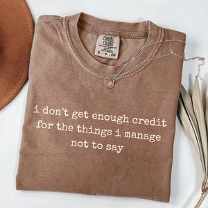 May include: A brown short-sleeved t-shirt with a crew neckline and a Comfort Colors brand label. The shirt features white printed text that reads: i don't get enough credit for the things i manage not to say. A silver chain necklace rests on top.
