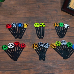 May include: Several black hair clips, each with a different coloured circular emblem. The emblems feature symbols such as a star, a skull, and geometric patterns. The clips are displayed on a dark wooden surface.