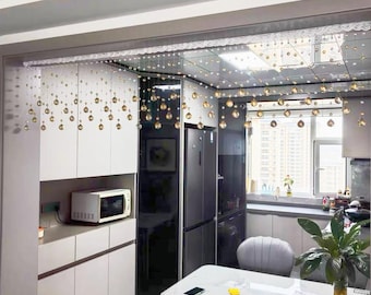 May include: A kitchen interior featuring a decorative beaded curtain with gold-colored orbs. The curtain hangs from a white trim above a kitchen area with white cabinets, a microwave, and a refrigerator. A table and chair are also visible.