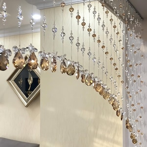 May include: A decorative crystal beaded curtain. The curtain features clear and amber-colored crystals of various shapes and sizes, strung together on thin wires. The crystals hang in a curved pattern, creating a shimmering effect.