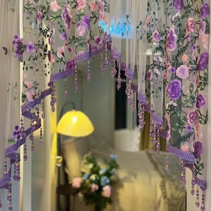 May include: Sheer floral curtains with embroidered purple and pink flowers. The curtains have a decorative purple ribbon trim with dangling beaded accents. The design creates a romantic and elegant aesthetic, suitable for a bedroom or living space.