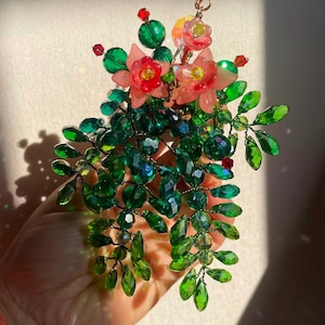 May include: A handmade floral accessory with green and clear crystal beads, and pink and red accents. The design includes delicate leaves and flowers, creating a sparkling, nature-inspired piece. The accessory is held in a hand.
