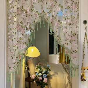 May include: Sheer floral curtain with pink flowers and green leaves. The curtain has a scalloped green trim and green tassel accents. The curtain is hanging in a doorway, with a glimpse of a bedroom interior.