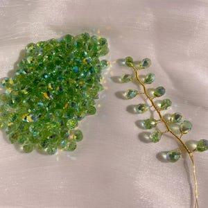 May include: A collection of green, iridescent crystal beads and a delicate gold wire floral stem. The beads are clustered together, while the stem features individual crystal droplets. The background is a soft, white fabric.