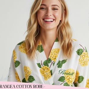 Puede incluir: Bata de algod&oacute;n blanca con un estampado floral de hortensias amarillas y hojas verdes. La bata tiene un dise&ntilde;o envolvente y el nombre "Kendall" est&aacute; bordado en escritura verde. El texto "~HYDRANGEA COTTON ROBE~" est&aacute; en la parte inferior.
