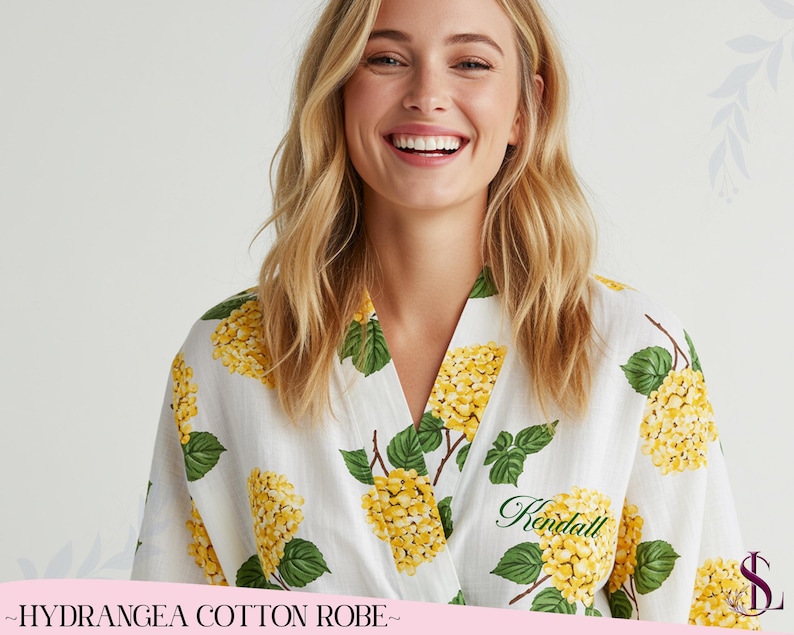 Puede incluir: Bata de algod&oacute;n blanca con un estampado floral de hortensias amarillas y hojas verdes. La bata tiene un dise&ntilde;o envolvente y el nombre "Kendall" est&aacute; bordado en escritura verde. El texto "~HYDRANGEA COTTON ROBE~" est&aacute; en la parte inferior.