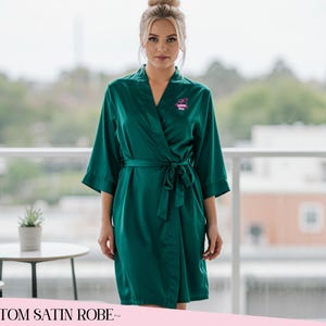 May include: A dark green satin robe with a wrap-around style and a tie belt. The robe features three-quarter length sleeves and a small embroidered logo. The text "CUSTOM SATIN ROBE" is displayed at the bottom of the image.