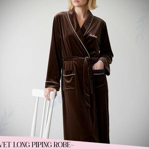Op de afbeelding: Een lange, bruine fluwelen badjas met witte biezen en een bijpassende ceintuur. De badjas heeft zakken en de naam "Andrea" geborduurd op de borst. De badjas maakt deel uit van een loungewear collectie.