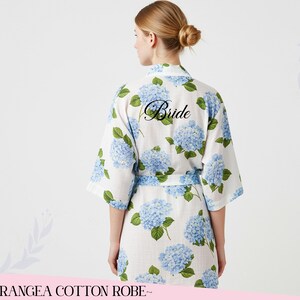 Puede incluir: Bata de algod&oacute;n blanca con un estampado floral de hortensias azules. La palabra "Bride" est&aacute; impresa en la espalda en escritura negra. La bata tiene un cintur&oacute;n a juego y mangas cortas. El texto "~HYDRANGEA COTTON ROBE~" est&aacute; impreso en la parte inferior.