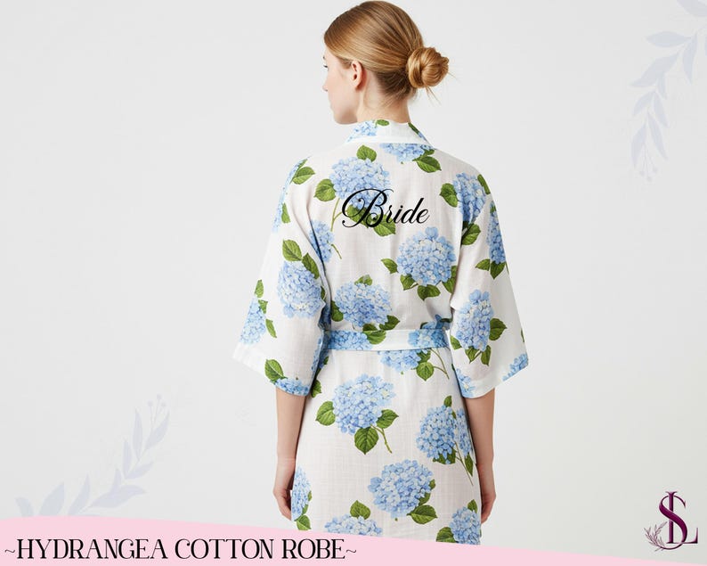 Puede incluir: Bata de algod&oacute;n blanca con un estampado floral de hortensias azules. La palabra "Bride" est&aacute; impresa en la espalda en escritura negra. La bata tiene un cintur&oacute;n a juego y mangas cortas. El texto "~HYDRANGEA COTTON ROBE~" est&aacute; impreso en la parte inferior.