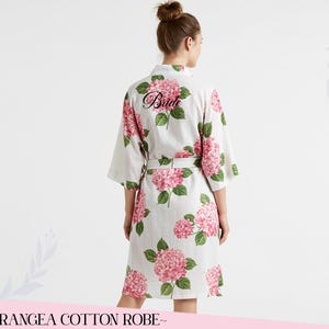 Puede incluir: Bata de algod&oacute;n blanca con un estampado floral de hortensias rosas. La palabra "Bride" est&aacute; impresa en la espalda en escritura negra. La bata tiene un cintur&oacute;n a juego y mangas tres cuartos. El texto "~HYDRANGEA COTTON ROBE~" est&aacute; en la parte inferior.