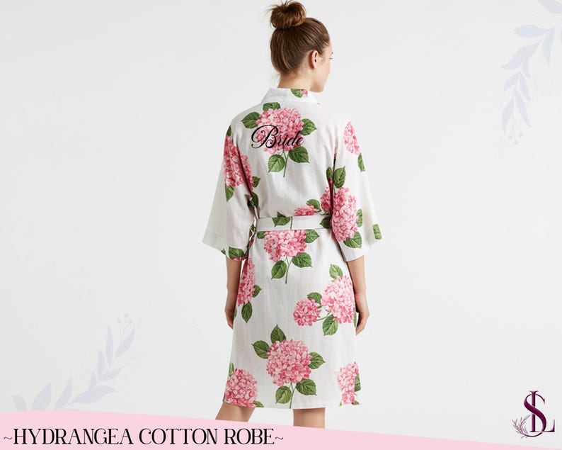 Puede incluir: Bata de algod&oacute;n blanca con un estampado floral de hortensias rosas. La palabra "Bride" est&aacute; impresa en la espalda en escritura negra. La bata tiene un cintur&oacute;n a juego y mangas tres cuartos. El texto "~HYDRANGEA COTTON ROBE~" est&aacute; en la parte inferior.