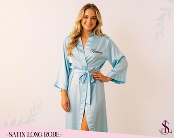 May include: A light blue satin long robe with a matching tie belt. The robe has wide sleeves and the name "Brittany" is embroidered on the chest in black script. The text "SATIN LONG ROBE" is at the bottom.