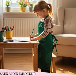 Puede incluir: Una niña con un delantal verde con la palabra "Chef" bordada, dibuja en una pequeña mesa de madera. Lápices de colores en tazas verdes y amarillas. La niña lleva una camisa a rayas.