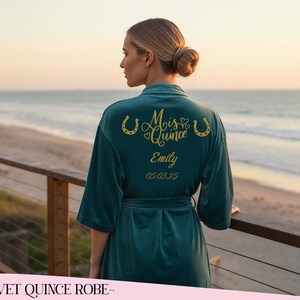 May include: A dark teal velvet robe featuring gold lettering on the back that reads Mis Quince, the name Emily, and the date 05.03.25. The design includes two gold horseshoe icons and small heart shapes flanking the text Mis Quince.
