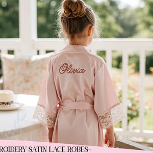 Könnte beinhalten: Hellrosa Satin-Bademantel mit Spitzenbesatz an den Ärmeln. Der Name "Olivia" ist in brauner Schrift auf den Rücken gestickt. Der Bademantel hat einen passenden Gürtel und ist Teil einer Kollektion von bestickten Satin-Spitzen-Roben.