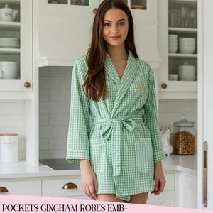 May include: A green and white gingham robe with a matching tie belt. The robe has a shawl collar, long sleeves, and two front pockets. The robe is knee-length and has white trim. The word "Grace" is embroidered on the chest.