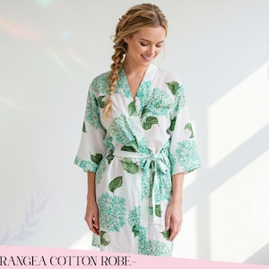 Puede incluir: Bata de algod&oacute;n blanca con un estampado floral de hortensias azules y verdes. La bata tiene un cierre de lazo y la palabra "Bride" bordada en la parte delantera. Parte de la colecci&oacute;n "Hydrangea Cotton Robe".