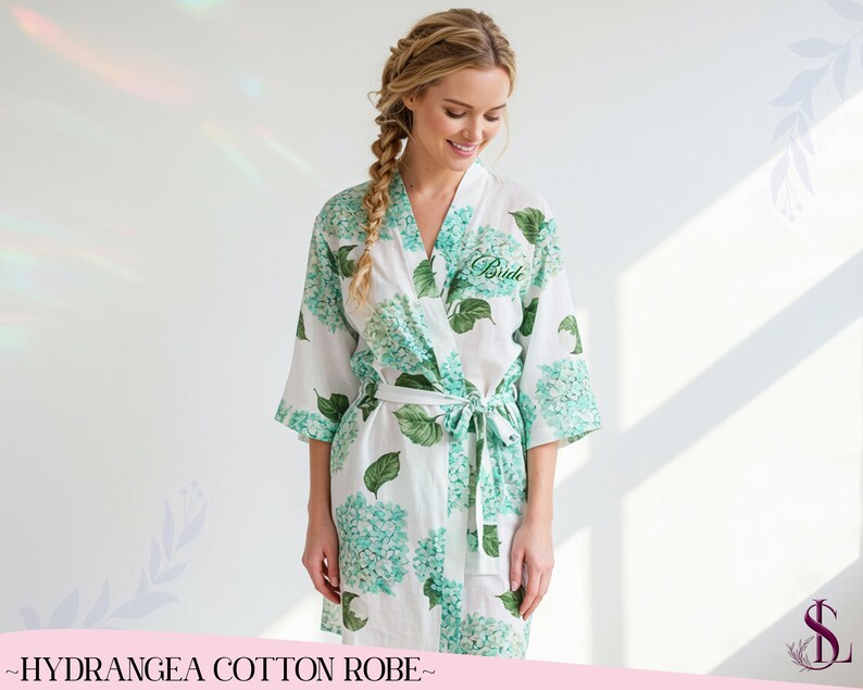 Puede incluir: Bata de algod&oacute;n blanca con un estampado floral de hortensias azules y verdes. La bata tiene un cierre de lazo y la palabra "Bride" bordada en la parte delantera. Parte de la colecci&oacute;n "Hydrangea Cotton Robe".
