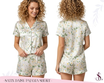 May include: A satin pajama short set featuring a daisy floral pattern. The set comprises a short-sleeved, button-down top and matching shorts. The fabric is white with yellow and green floral details, edged with green piping. The text at the bottom reads "SATIN DAISY PAJAMA SHORT".