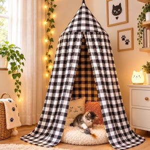Op de afbeelding: Een conische hangende baldakijntent met een zwart-wit ruitpatroon. Binnenin de tent zit een klein kitten op een rond, crèmekleurig pluche kussen naast een wit kussen in de vorm van een kat en een roze kussen met textuur.