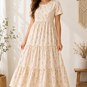 May include: A cream-coloured midi dress featuring a delicate brown and orange floral print, short sleeves, a round neckline, and a tiered skirt design that falls to the mid-calf. The dress is paired with simple beige strappy sandals.