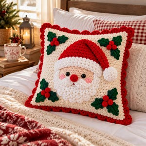 Puede incluir: Un cojín cuadrado de ganchillo con una cara de Papá Noel en el centro, con gorro rojo, barba blanca y mejillas rosadas. El fondo color crema está decorado con cuatro grupos de hojas de acebo verdes y bayas rojas, con un borde festoneado rojo.