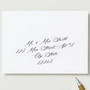 Puede incluir: Un sobre blanco con elegante caligrafía negra. El texto incluye "Mr. & Mrs. Smith", una dirección y un código postal. Un bolígrafo con empuñadura dorada descansa en la esquina inferior derecha.
