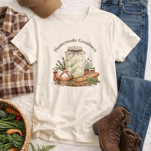 Peut inclure: T-shirt crème avec l'inscription "Homemade Goodness" et un dessin d'un bocal, de pain et d'herbes. L'image comprend également une casquette de baseball marron, une chemise à carreaux, un jean bleu et des bottes marron.