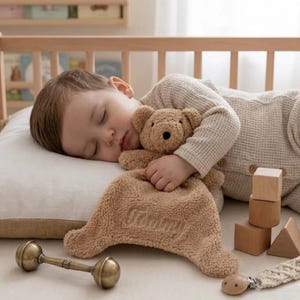 Puede incluir: Un niño dormido abraza una mantita de seguridad con forma de oso de peluche marrón con el nombre "Tommy" bordado. El niño lleva una camisa de punto de gofre beige y está acostado sobre una almohada blanca. Un sonajero vintage y bloques de madera están cerca.