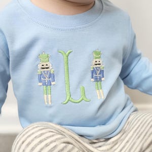 May include: Light blue long-sleeved shirt with embroidered nutcracker soldiers and a green letter "L". The shirt is paired with gray and white striped pants. The design is suitable for children.