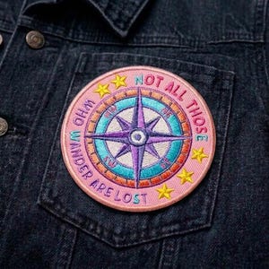 May include: Embroidered patch with a compass design in shades of purple, teal, and pink, with the text "NOT ALL THOSE WHO WANDER ARE LOST." The patch is circular with yellow star accents and is attached to a dark denim jacket.