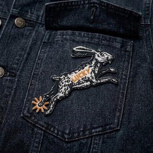 May include: Close-up of a dark denim jacket with a pocket. A patch of a running rabbit with the word "LUCKY" in orange is sewn onto the pocket. The rabbit is black and white with a gold spur.