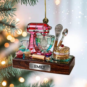 May include: A decorative Christmas ornament featuring a pink and multi-colored stand mixer, cupcakes, and a cake. The ornament includes a rolling pin, whisk, and spoon. The name "EMILY" is on a silver plaque on the wooden base.
