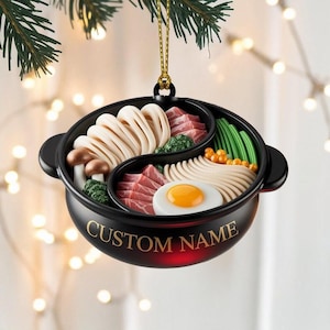 May include: A black ornament shaped like a bowl of ramen, with a gold hanging string. The bowl is divided into two sections, filled with noodles, meat, vegetables, and an egg. The text "CUSTOM NAME" is printed on the bowl.
