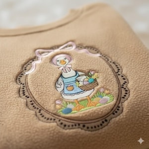 May include: Tan fleece fabric with an embroidered design. The design features a goose wearing a bonnet and apron, holding a basket of Easter eggs. The embroidery is framed by a scalloped border with a pink ribbon.